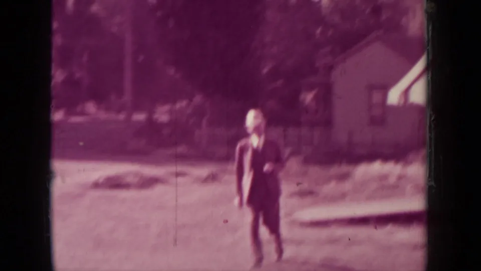 The man in a suit and hat moves closer to the camera, with a house and storefront awning behind him.