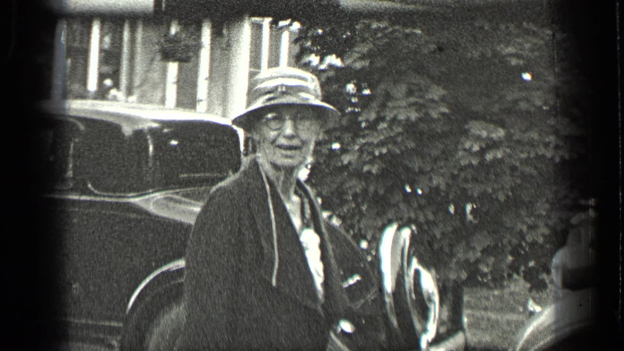 Older woman wearing glasses, a dark coat, and a brimmed hat stands near vintage cars in Long Lake, Canada, 1939.