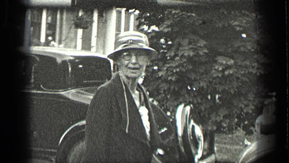 Older woman wearing glasses, a dark coat, and a brimmed hat stands near vintage cars in Long Lake, Canada, 1939.