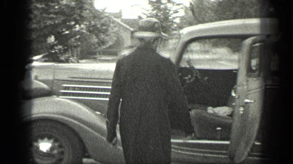 The woman reaches the open door of a grey vintage car, with another parked vehicle and trees visible nearby.