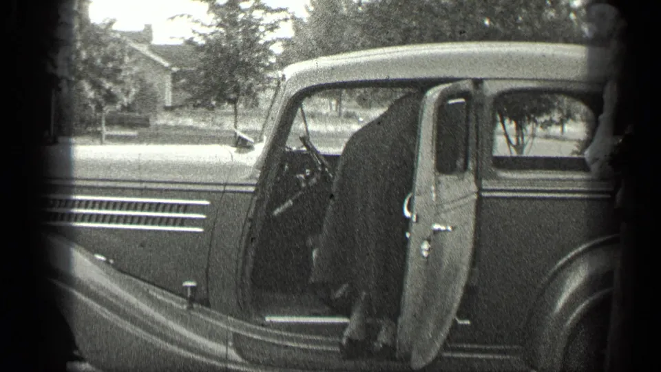 In Long Lake, Canada, a rounded late-1930s car sits with its door open near a house and trees.