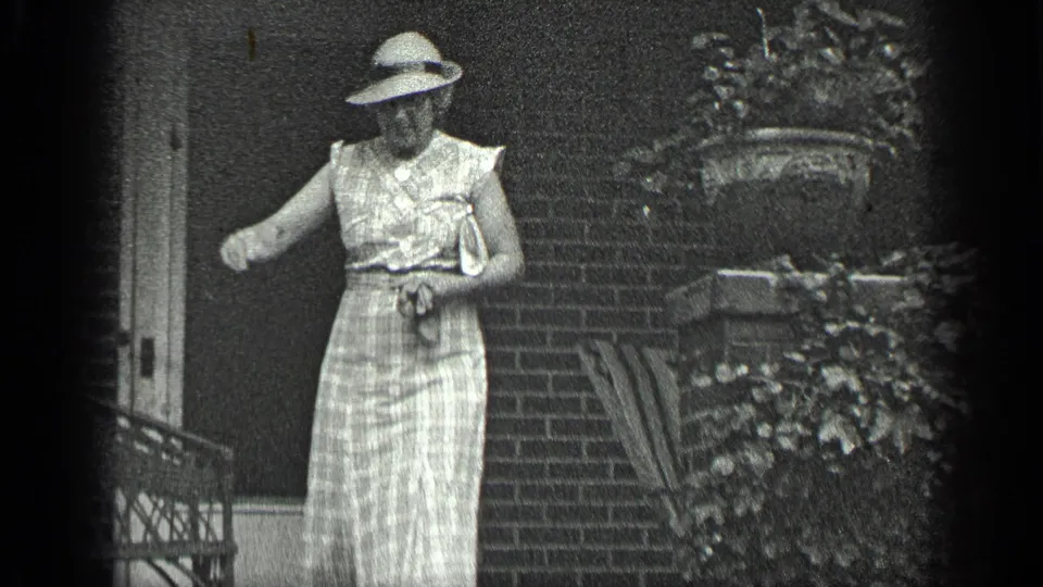 A woman in a light dress and wide-brimmed hat stands by the brick porch steps in the 1939 Long Lake footage.