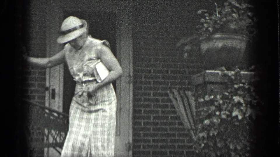 The woman descends beside the brick house entrance, her face partly shaded by a wide-brimmed hat.