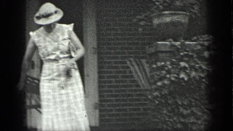 The woman turns toward the doorway at the brick house, reaching near the frame as the shot ends.