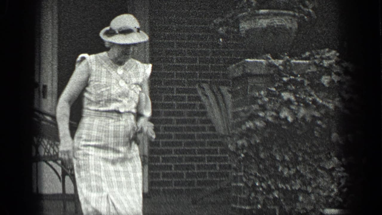 In Long Lake, Canada, 1939, a woman in a sleeveless checked dress and hat stands outside a brick house near steps and foliage.