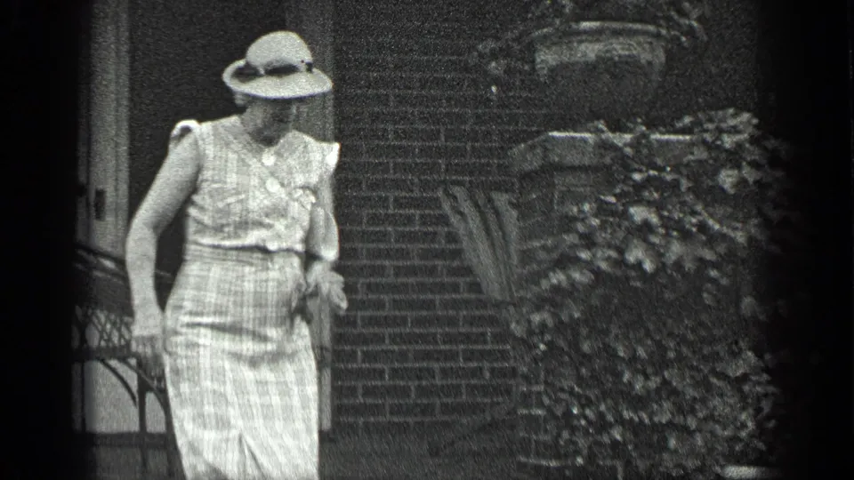 In Long Lake, Canada, 1939, a woman in a sleeveless checked dress and hat stands outside a brick house near steps and foliage.