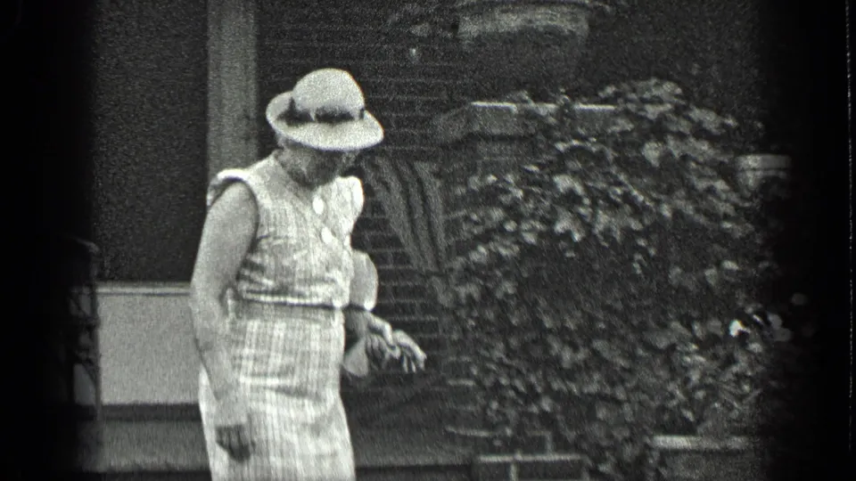 The woman walks past the brick entrance and garden foliage, wearing a wide-brimmed hat and sleeveless checked dress.