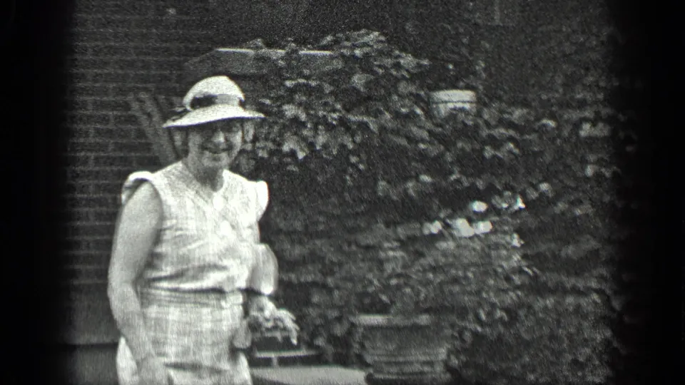 A smiling woman in 1939 home-movie footage stands near the garden edge, with brickwork and plants behind her.