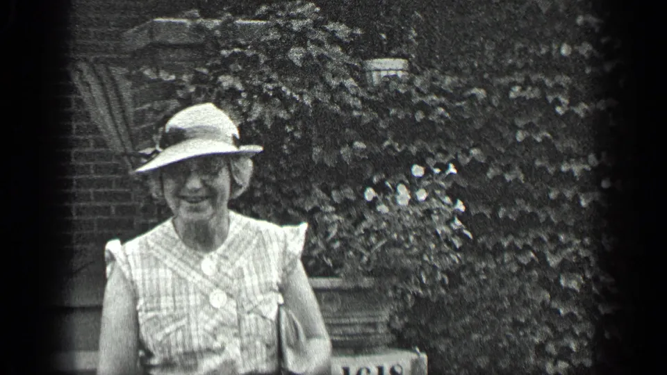 A closer Long Lake home-movie frame shows the woman smiling toward the camera in front of brickwork and leafy plants.