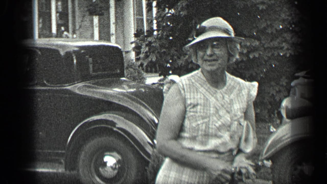 Older woman in a hat faces the camera between parked classic cars.