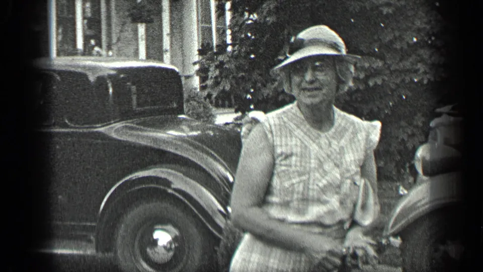 Older woman in a wide-brimmed hat faces the camera near parked classic cars during the 1939 Long Lake home movie.