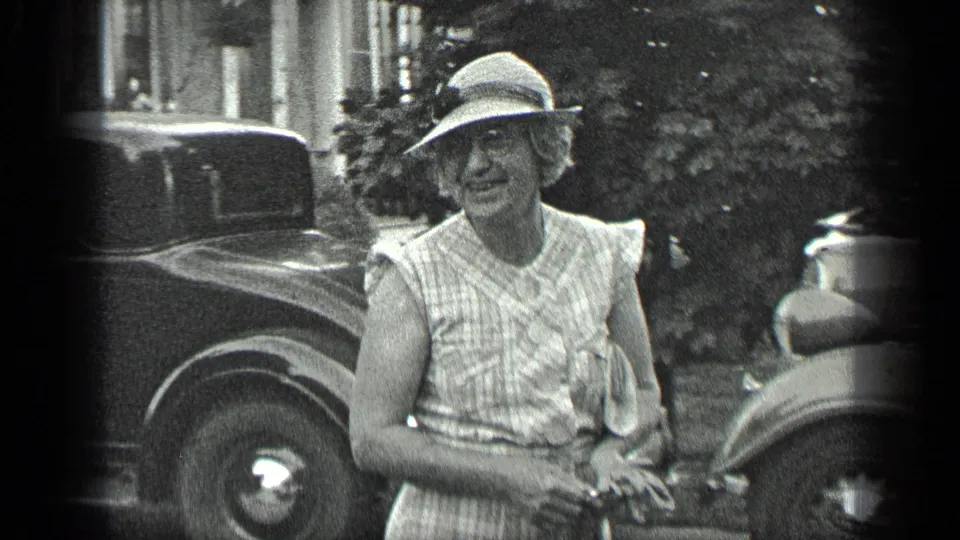 Older woman smiles near classic automobiles in a residential outdoor setting in the Long Lake, Canada 1939 footage.