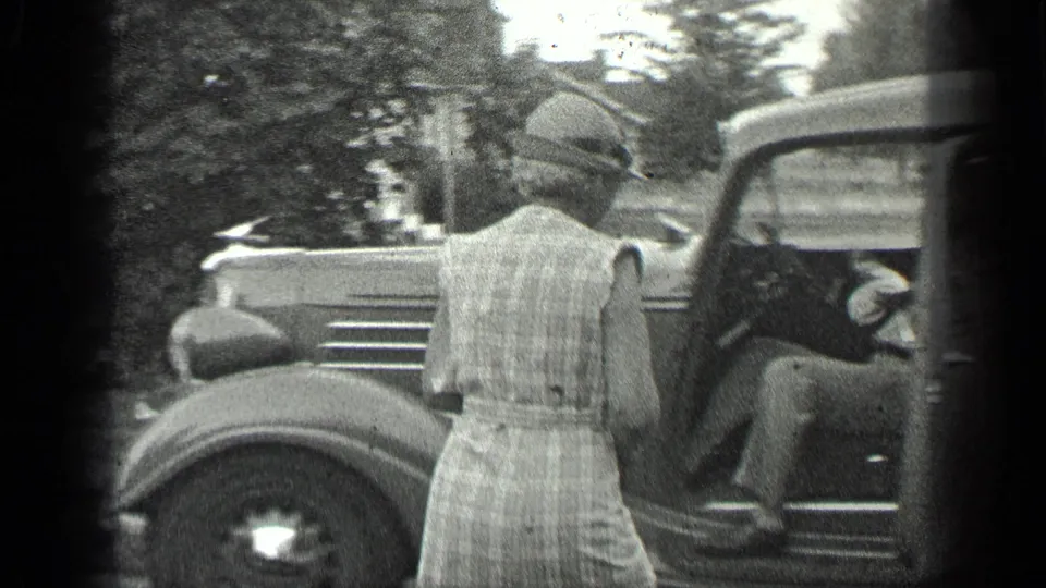 Woman in a hat stands at an open classic car door as a seated person is visible inside, likely preparing to enter the vehicle.