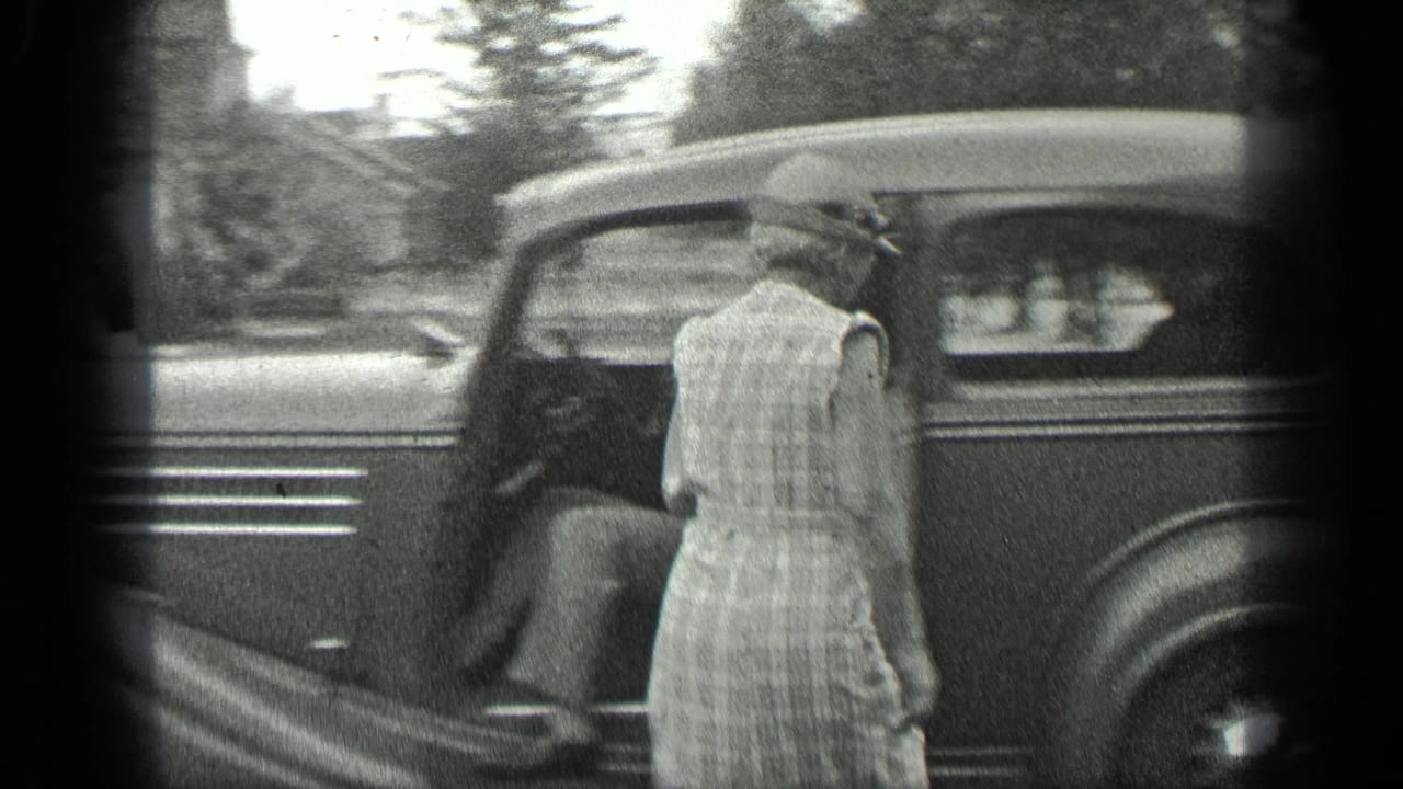 Woman in a light dress and hat stands beside a dark vintage car near trees.