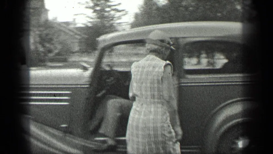 In 1939 Long Lake, Canada footage, a woman in a light dress and hat stands by the open side of a dark vintage car.