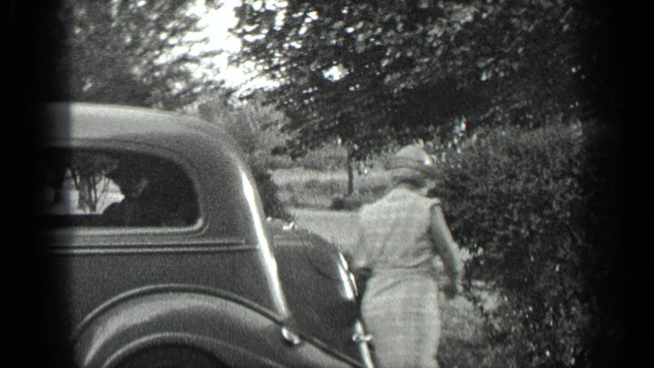 Woman in a light dress walks away from the rear of a dark vintage car.