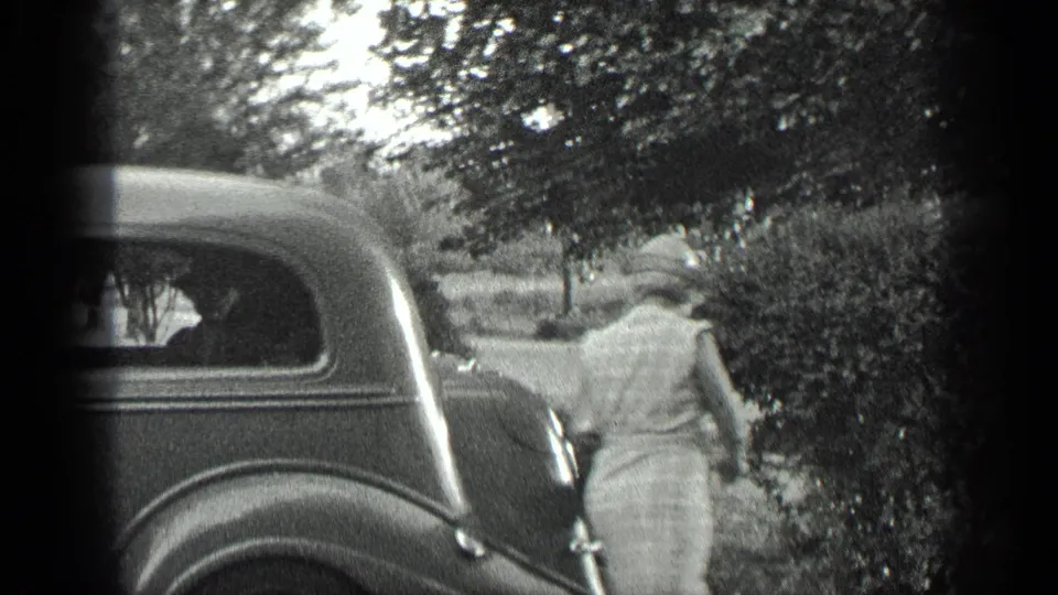 A woman in a light dress and hat walks near the rear of a dark vintage car along a tree-lined roadside in Long Lake, Canada.