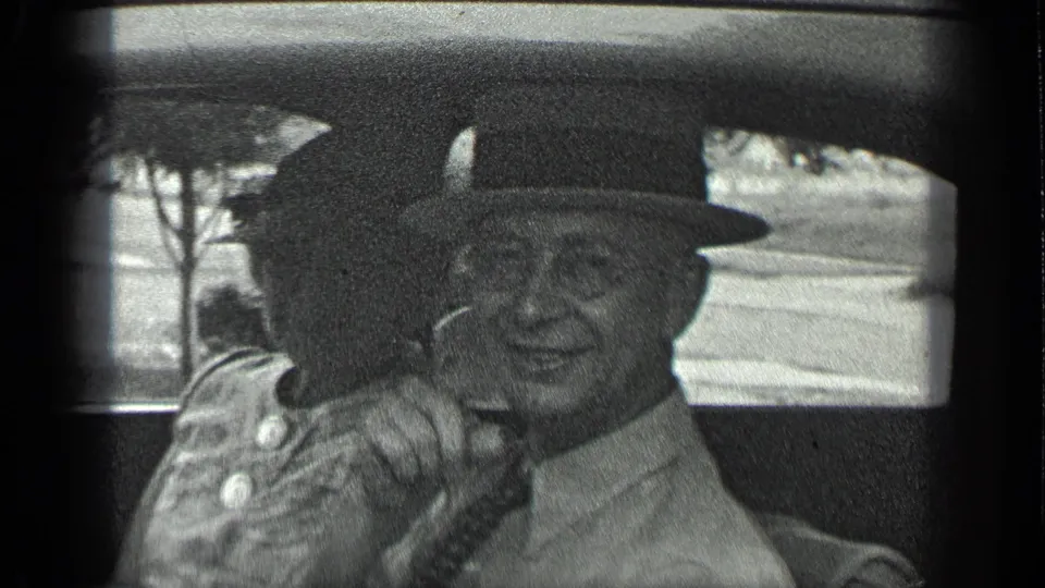 An elderly couple sits inside the car; the man in a fedora and glasses smiles toward the camera while a woman sits beside him.