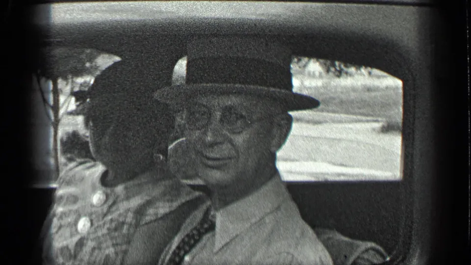 The older man in fedora and glasses looks toward the camera beside the woman in the 1939 Long Lake, Canada car footage.
