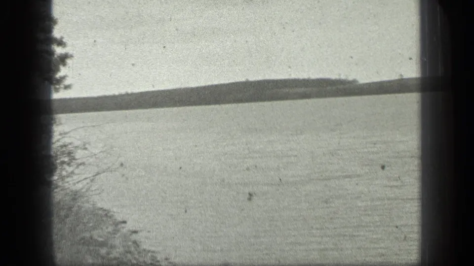 A handheld 8mm view shows Long Lake water with shoreline vegetation in the foreground and low wooded land beyond.