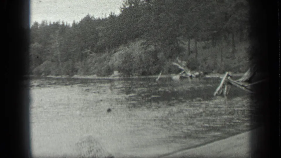 1939 Long Lake, Canada home-movie frame showing calm water, driftwood near shore, and dense trees across the river.