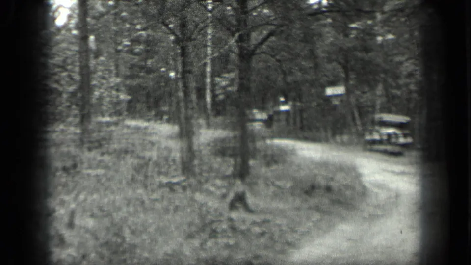 Black-and-white 1939 Long Lake frame with a dirt road, wooded verge, roadside signs, and a vintage car in the distance.