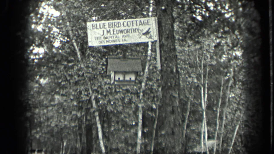 Home-movie view of the Blue Bird Cottage sign with J.M. Edworthy lettering, a birdhouse below, and surrounding woodland.