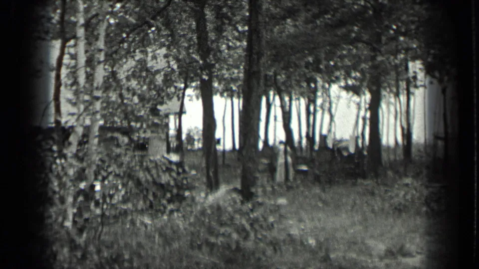 1939 Long Lake home-movie frame with a wooded clearing and a distant woman partly obscured among trees near a rustic structure.
