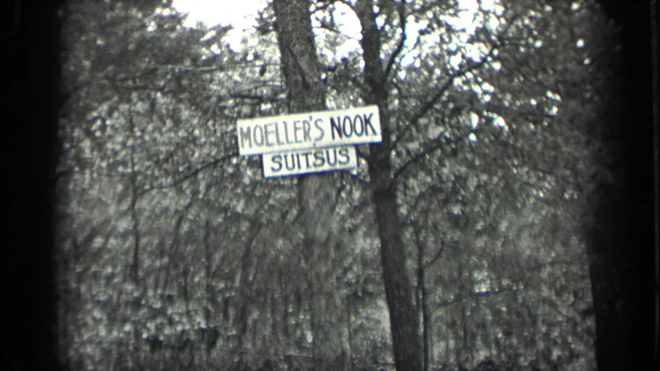 Final 1939 Long Lake frame holding on the Moeller's Nook Suitsus sign near a tree, with dense woodland behind.