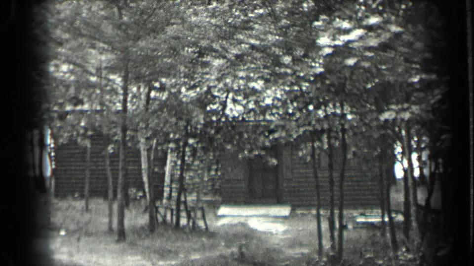 Black-and-white 8mm still from Long Lake showing a log cabin, doorway, trees, and a bench in a wooded clearing.
