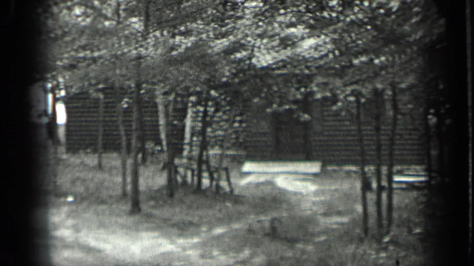 Late-1930s Long Lake frame showing a rustic log cabin behind trees, with a pale bench and wooded ground in the foreground.