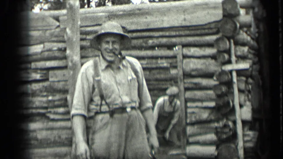 1939 Long Lake, Canada frame with a man in suspenders and a broad hat standing in front of a rustic log cabin.