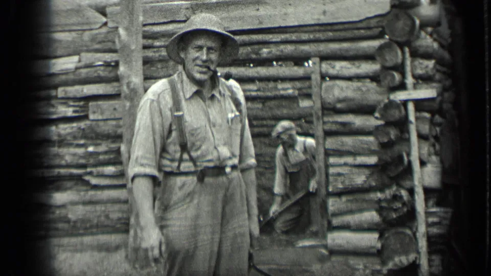 A man in work clothes and suspenders stands before a log cabin at Long Lake, Canada, with another person near the doorway.