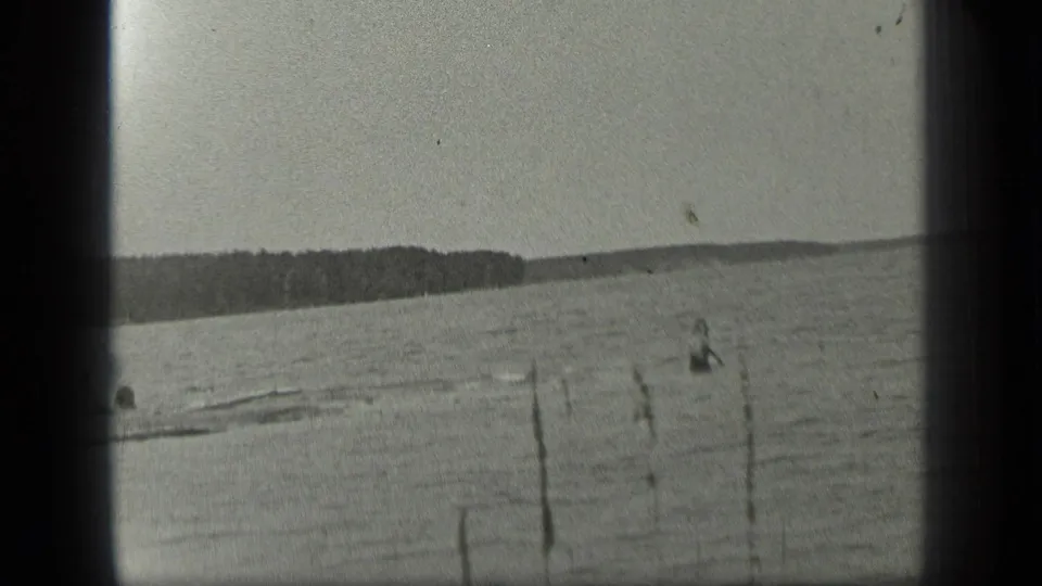 Wide view across Long Lake with distant swimmers, shoreline posts, and a dark treeline in 1939 home-movie footage.