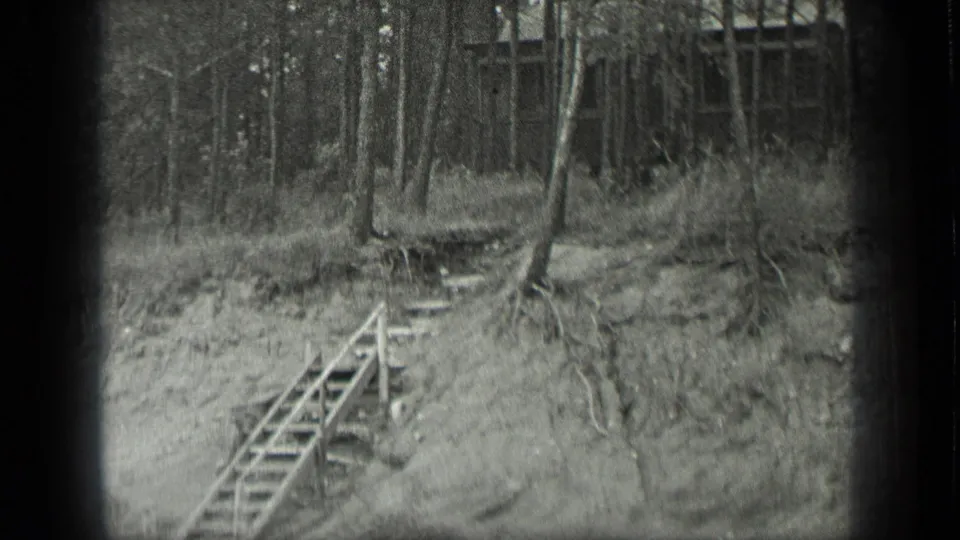 1939 archival home-movie frame showing wooden steps, exposed roots, trees, and a cabin set back in the Long Lake woods.