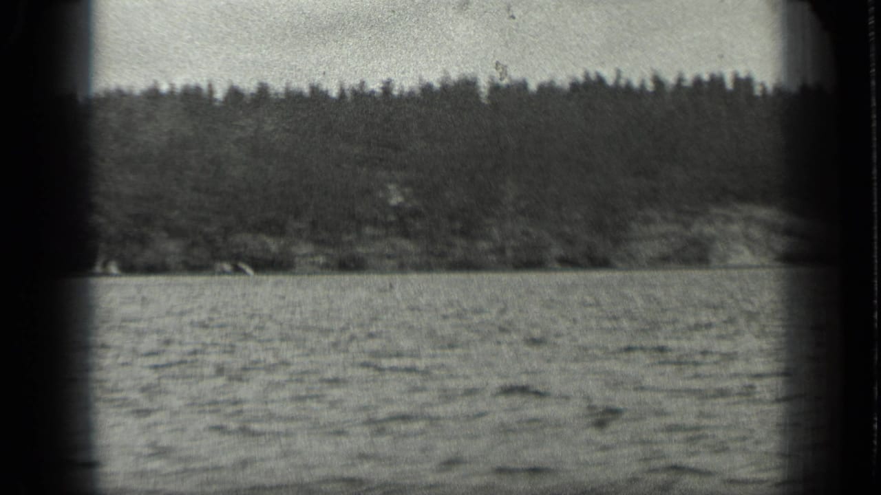 1939 Long Lake, Canada frame with rippled water in the foreground and dense forest along the far shore, seen through soft 8mm grain.