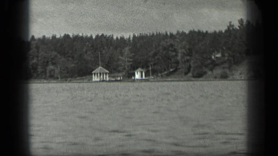 1939 8mm view from Long Lake, Canada, looking across rippled water toward a wooded shore with two small white lakeside pavilions or cabins.