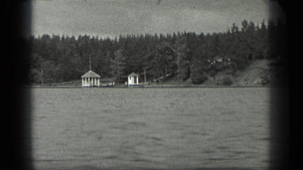 Lake surface and wooded shore with two small white buildings near the waterline.
