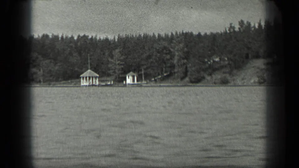 Boat-level 1939 Long Lake, Canada view with gentle water texture, dense trees, and two small light-colored shoreline structures.
