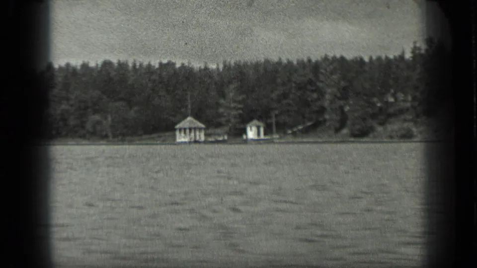 Soft 8mm frame from Long Lake, Canada, showing a forested bank, rippled water, and two small white structures along the shore.