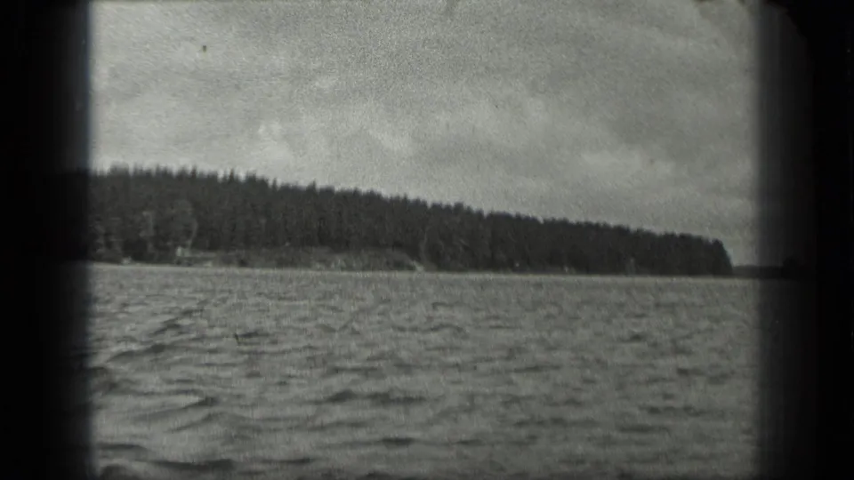 Boat-view 1939 Long Lake, Canada frame with rippled water in front of a continuous wooded shoreline and overcast sky.