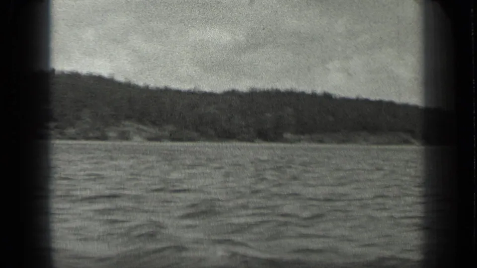 Black-and-white 8mm frame from Long Lake, Canada, showing open water and a low forested shoreline from a moving boat.