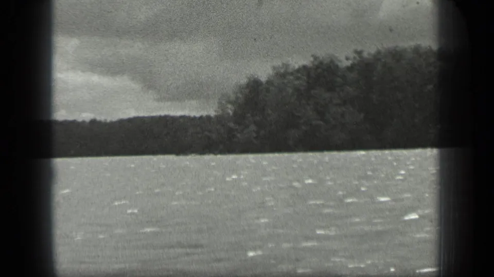 1939 boat-view frame on Long Lake, Canada, with bright ripples on the water and a wooded shoreline entering from the right.