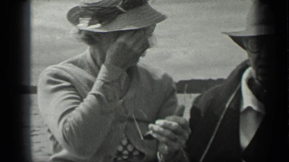 1939 Long Lake home-movie frame showing a woman adjusting eyewear beside a man in a fedora and glasses on a boat.