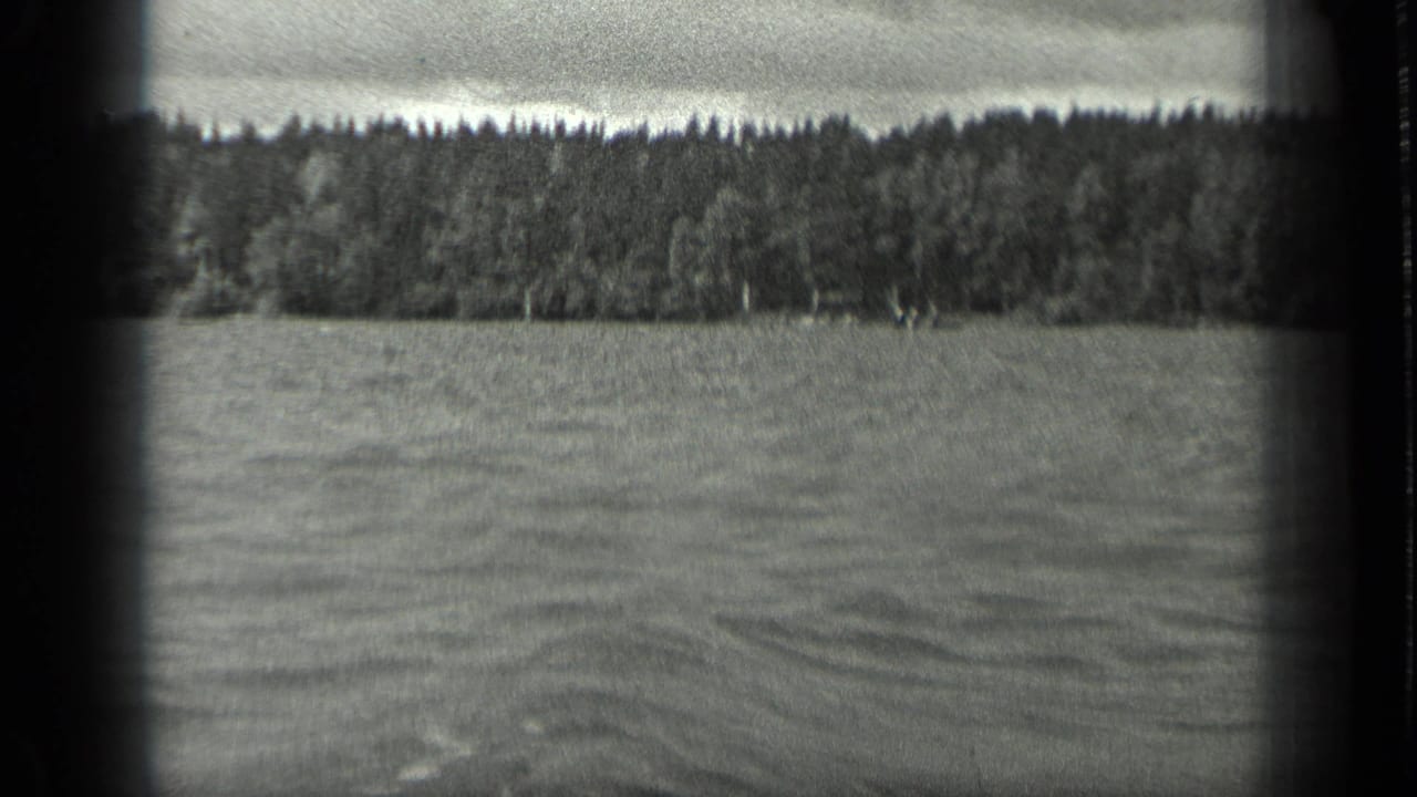 Black-and-white 8mm frame of Long Lake, Canada, showing rippled water and a dense wooded shore in the distance.
