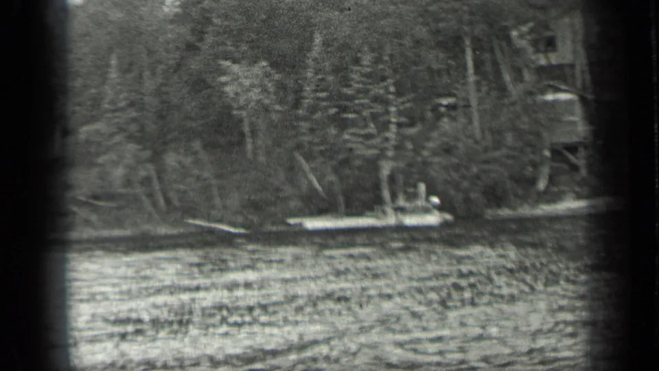 1939 Long Lake, Canada frame with a small white boat, wooden dock, rippled water, and a wooded lakeshore in soft 8mm focus.