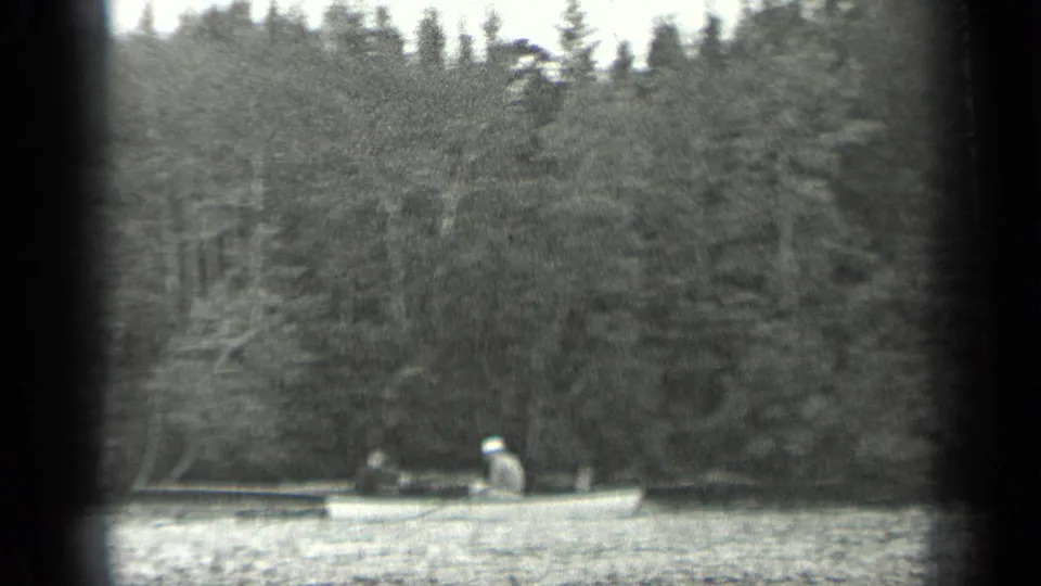 1939 Long Lake, Canada frame with an adult man in a light hat rowing a small boat across calm water before a wooded shoreline.