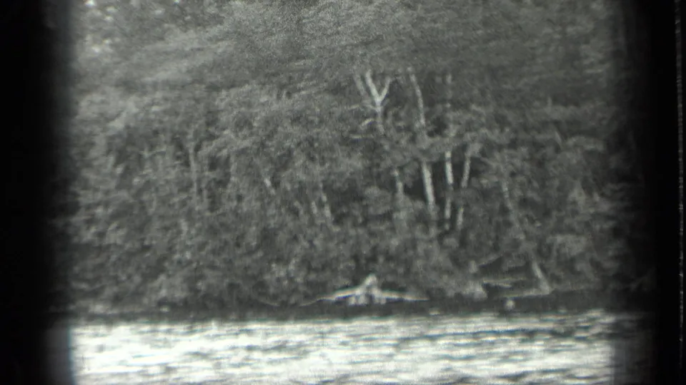 1939 Long Lake, Canada frame with a distant small boat low on the water beneath a thick forested shoreline.