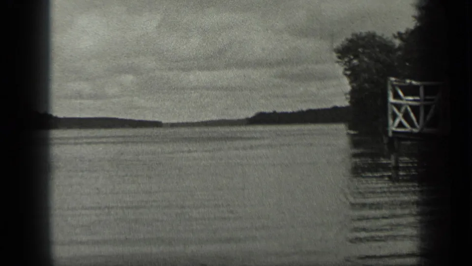 1939 Long Lake, Canada frame with calm water, distant shoreline, trees, and a white wooden dock or platform along the right edge.
