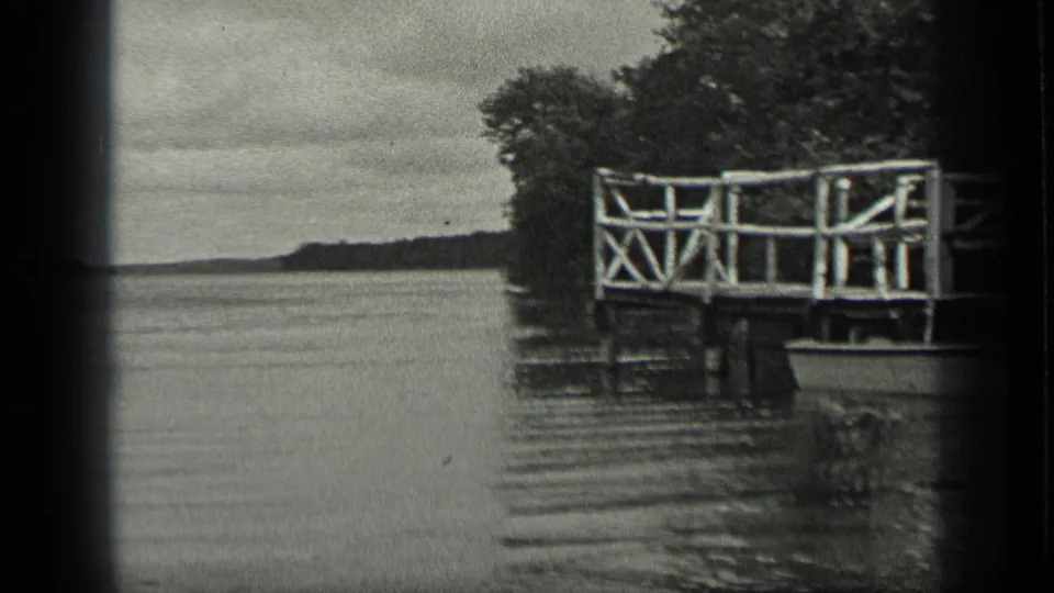 Black-and-white 8mm frame from Long Lake, Canada, showing a white wooden dock over calm water with trees along the shore.